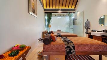 A modern hotel room with two beds, a wooden bench, a small table with flowers, a bathroom with a sink and mirror, and a large window.