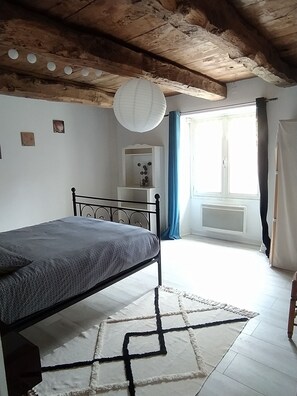 Quarto com uma cama, um radiador branco, uma janela com cortinas azuis e teto de madeira.
