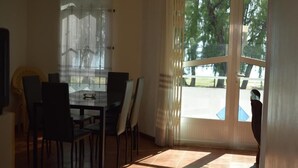 A dining area with a table and chairs, a window with curtains, and a view of trees outside. - Seafront Apartment Villa (Flic en Flac)