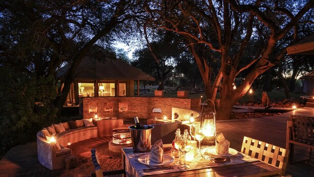 An outdoor dining area with a stone wall, wooden furniture, and ambient lighting. - Koro River Camp  (Tuli Lodge)