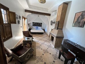 A bedroom with a bed, a chair, a small table, a decorative wall, and a fireplace. - Oloft (Marrakech)