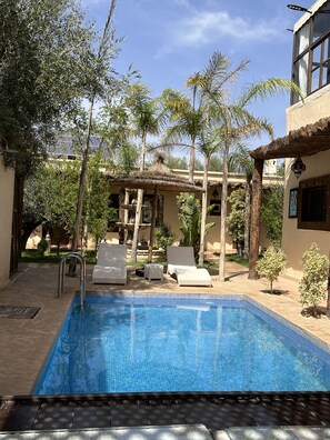 A swimming pool with lounge chairs, surrounded by palm trees and a building with a tiled roof. - Oloft (Marrakech)
