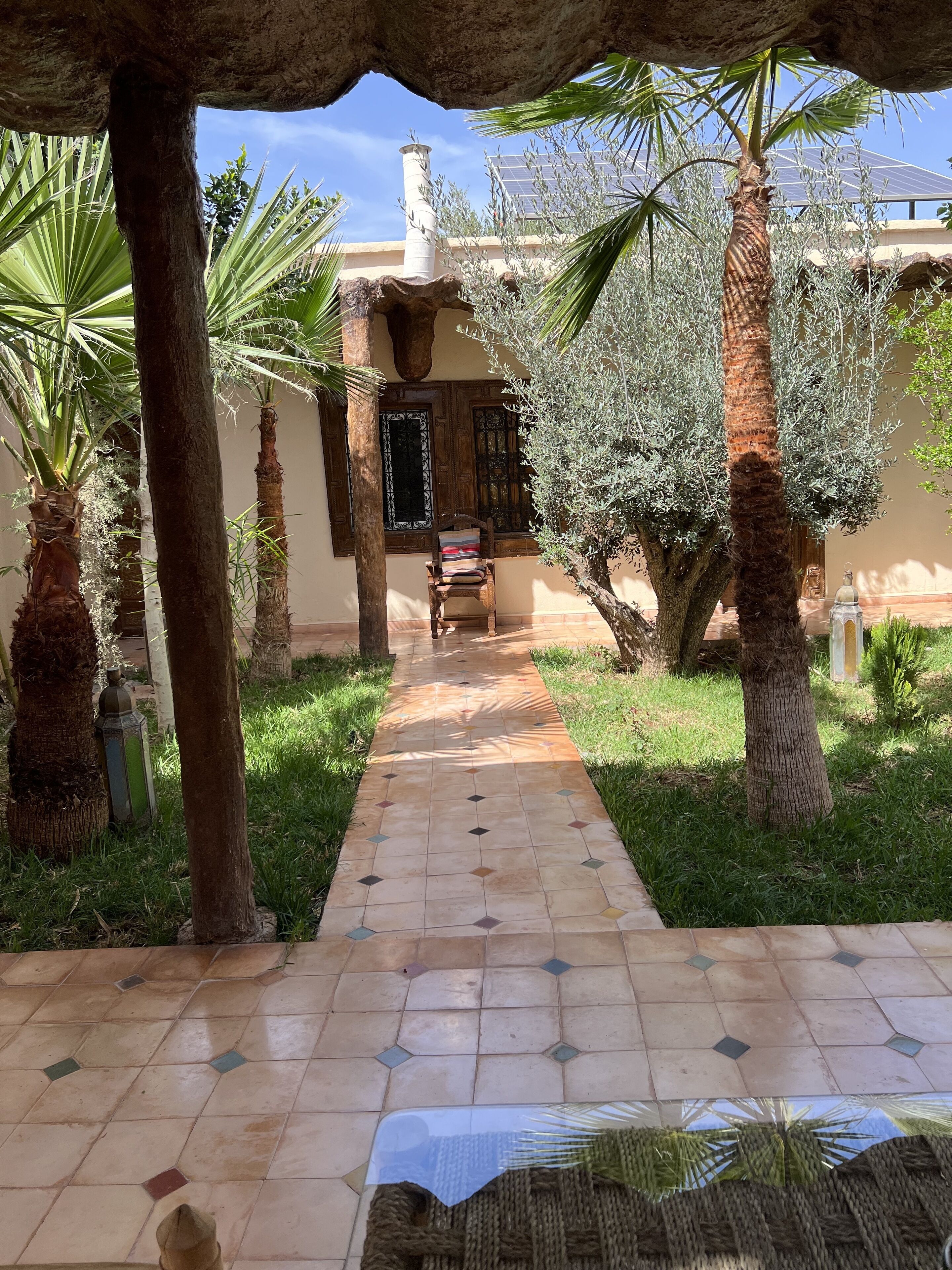 A pathway leading to a building with a tiled floor, surrounded by palm trees and a pergola structure.