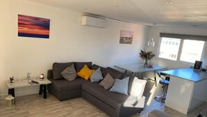 A modern living room with a grey sectional sofa, a small table with decorative items, a kitchen area with a white countertop, and a large window providing natural light. - Sea-side holiday home with balcony views to the sea (Hunstanton)