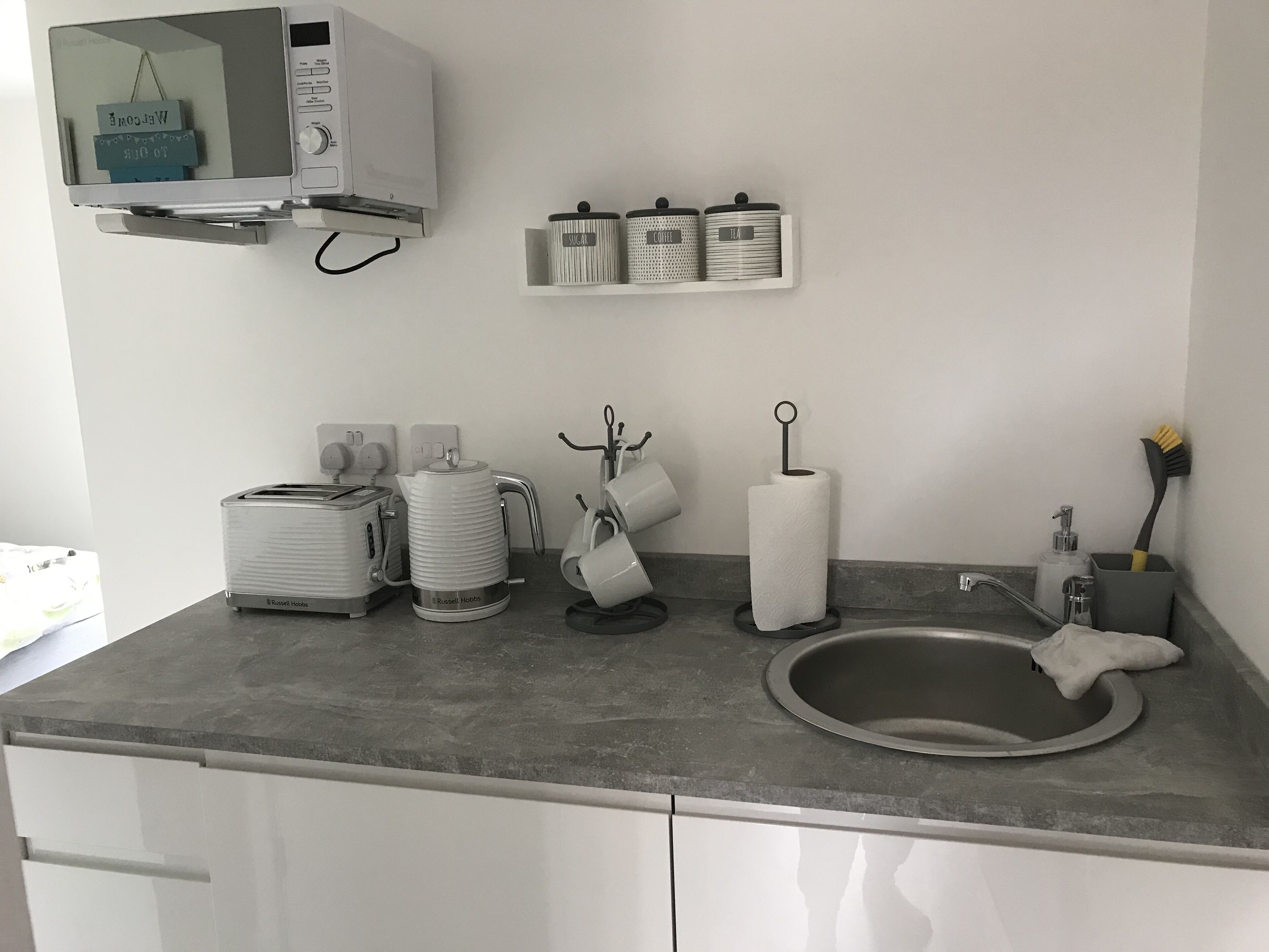 A kitchen with a built-in microwave, toaster, kettle, and sink. There are storage containers on the wall and a hanging towel rack.