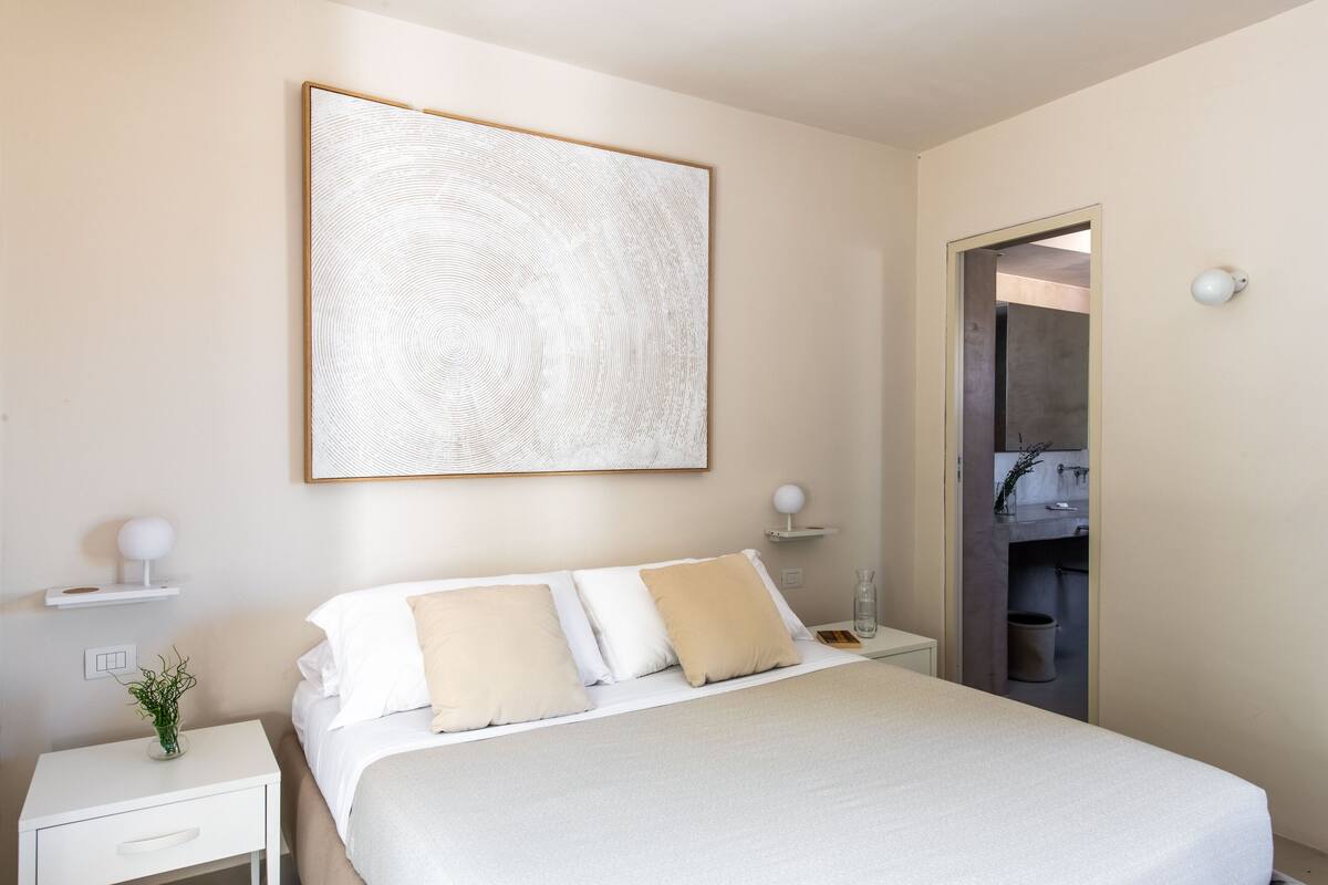 Une chambre à coucher comprenant un lit, une table de chevet, une lampe fixée au mur et une porte donnant sur une salle de bain.