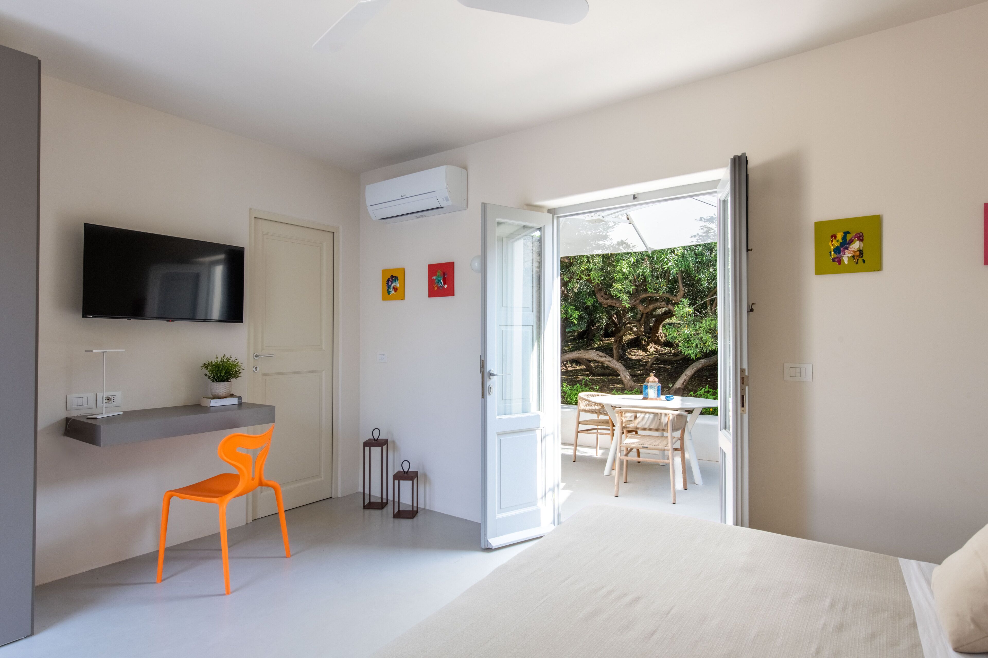 Un dormitorio con cama, escritorio con silla, televisor y puerta abierta que da a un balcón con mesa y sillas.