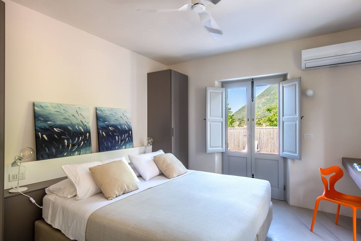 Une chambre à coucher avec un lit, deux tableaux, un bureau, une chaise orange et une vue sur l’extérieur par une porte ouverte.