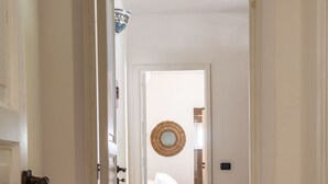 A narrow hallway leading to a bedroom with a bed, a round mirror on the wall, and a beige chair. - Vico San Lorenzo - Camere a Salina (Malfa)