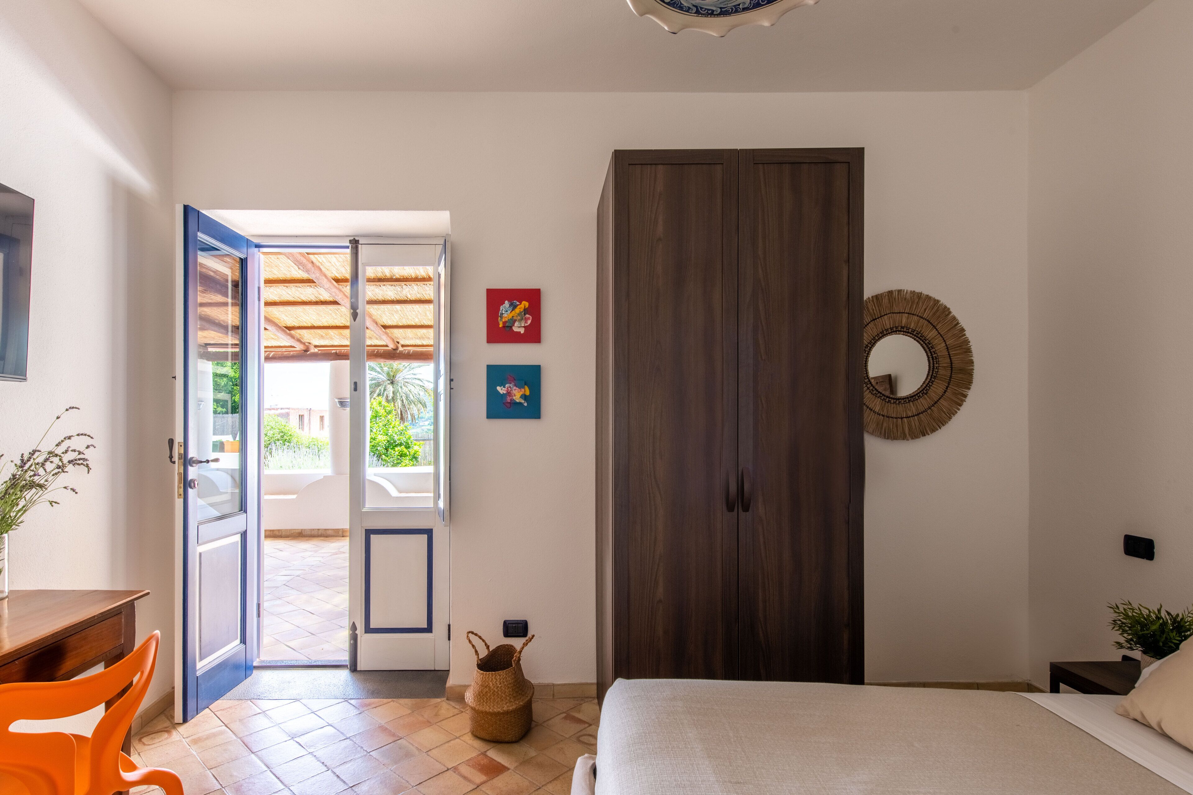Quarto com cama, armário, porta para uma varanda e uma pequena mesa com uma planta.