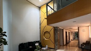 Um hall amplo com um sofá preto, um banquinho branco, uma mesa pequena com uma planta e um detalhe decorativo na parede.