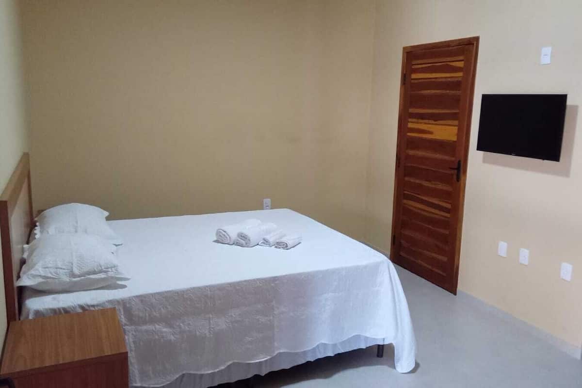 Una cama individual con sábanas blancas, cabecera de madera, una puerta de madera y un televisor de pantalla plana montado en la pared.