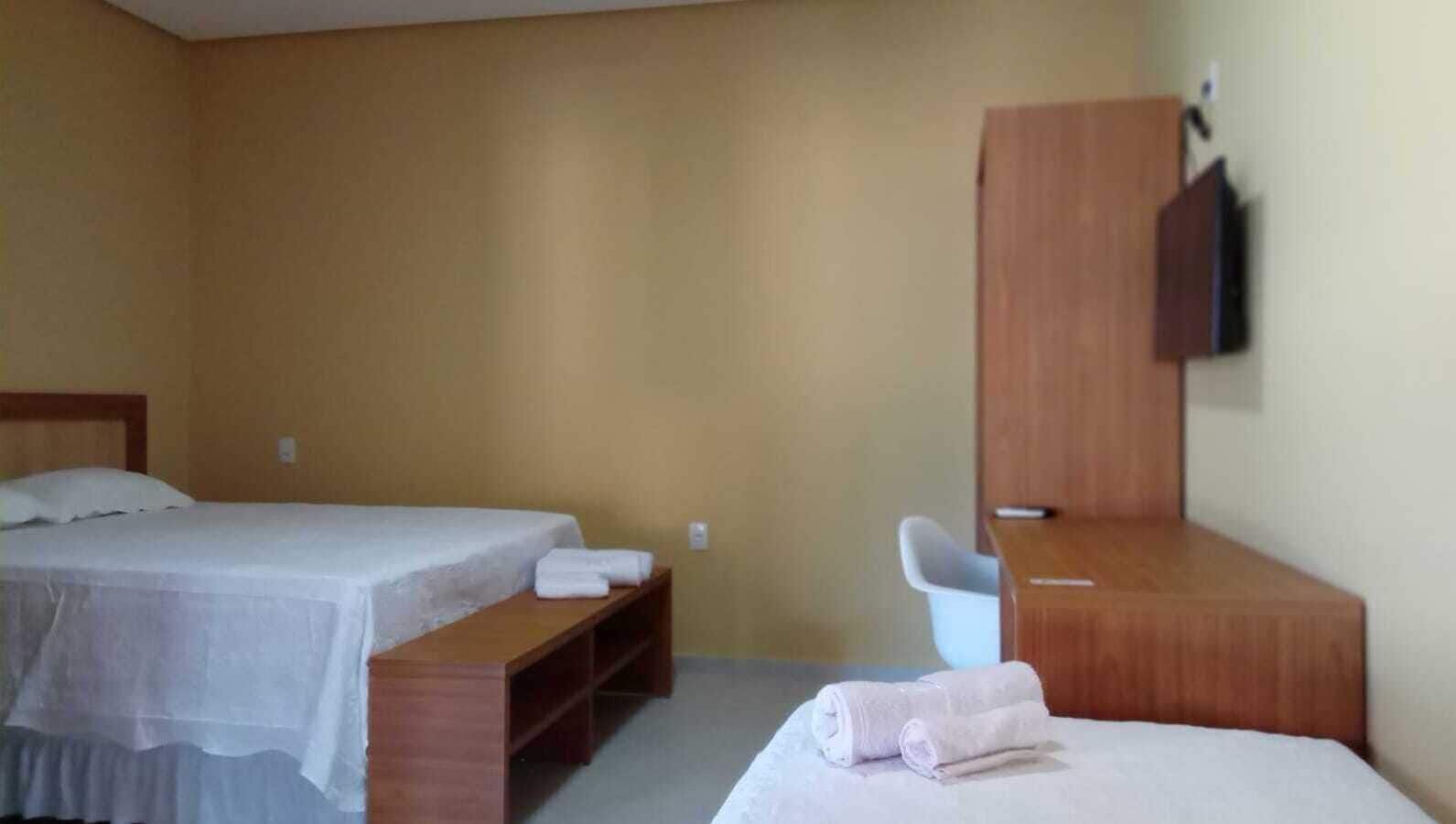 Quarto de hotel com duas camas, uma escrivaninha de madeira, uma cadeira e uma televisão fixada na parede.