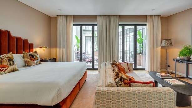Ein modernes Hotelzimmer mit einem großen Bett, einer Couch, einem Esstisch und Blick ins Freie durch große Fenster.