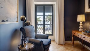 Een kamer met een donkerblauwe wand, een raam met witte gordijnen, een stoel, een tafel met een fles wijn en glas, en een lamp op een houten bijzettafeltje.