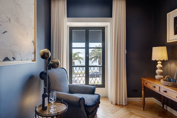 A room with a dark blue wall, a window with white curtains, a chair, a table with a wine bottle and glass, and a lamp on a wooden side table.