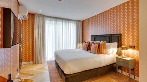 Una habitación de hotel con una cama grande, una mesita de noche con lámpara, un televisor montado en la pared y una ventana con cortinas.
