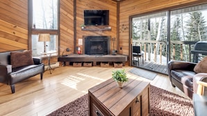 A cozy cabin interior with a fireplace, leather sofas, a television, and large windows offering a view of the forest. - Snowshoe Village 5A Ski In/Out Condo (Brian Head)