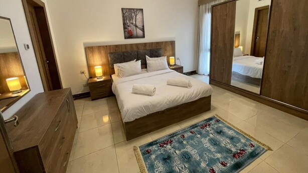 A bedroom with a large bed, bedside lamps, a wooden dresser, a mirror, and a blue patterned rug. - Luxury doublex apartments in new cairo
شقق فندقيه للايجار بالتجمع الخامس  (Cairo)