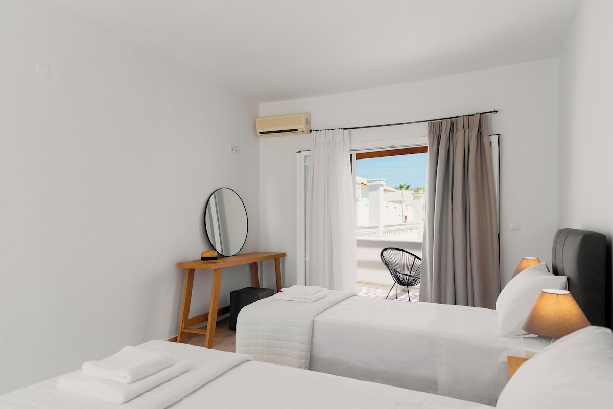 Une chambre d’hôtel avec deux lits, une table de chevet en bois, une chaise et une vue sur l’extérieur.