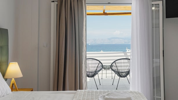 A hotel room with a bed, a nightstand with a lamp, and a view of the beach through a window with curtains.