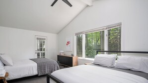 A bedroom with two beds, a window with a view of trees, a door, and a bedside table with a lamp. - Tate House (Saugatuck)