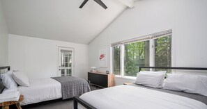 A bedroom with two beds, a window with a view of trees, a door, and a bedside table with a lamp. - Tate House (Saugatuck)