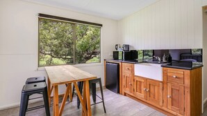 A compact kitchen with a wooden table and stools, a built-in sink, and a view of greenery through a large window.