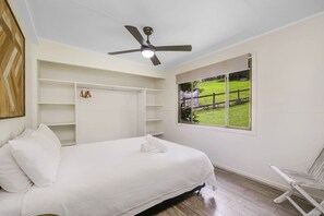 Une chambre à coucher avec un lit, un ventilateur de plafond, une fenêtre donnant sur un champ verdoyant et une chaise blanche.
