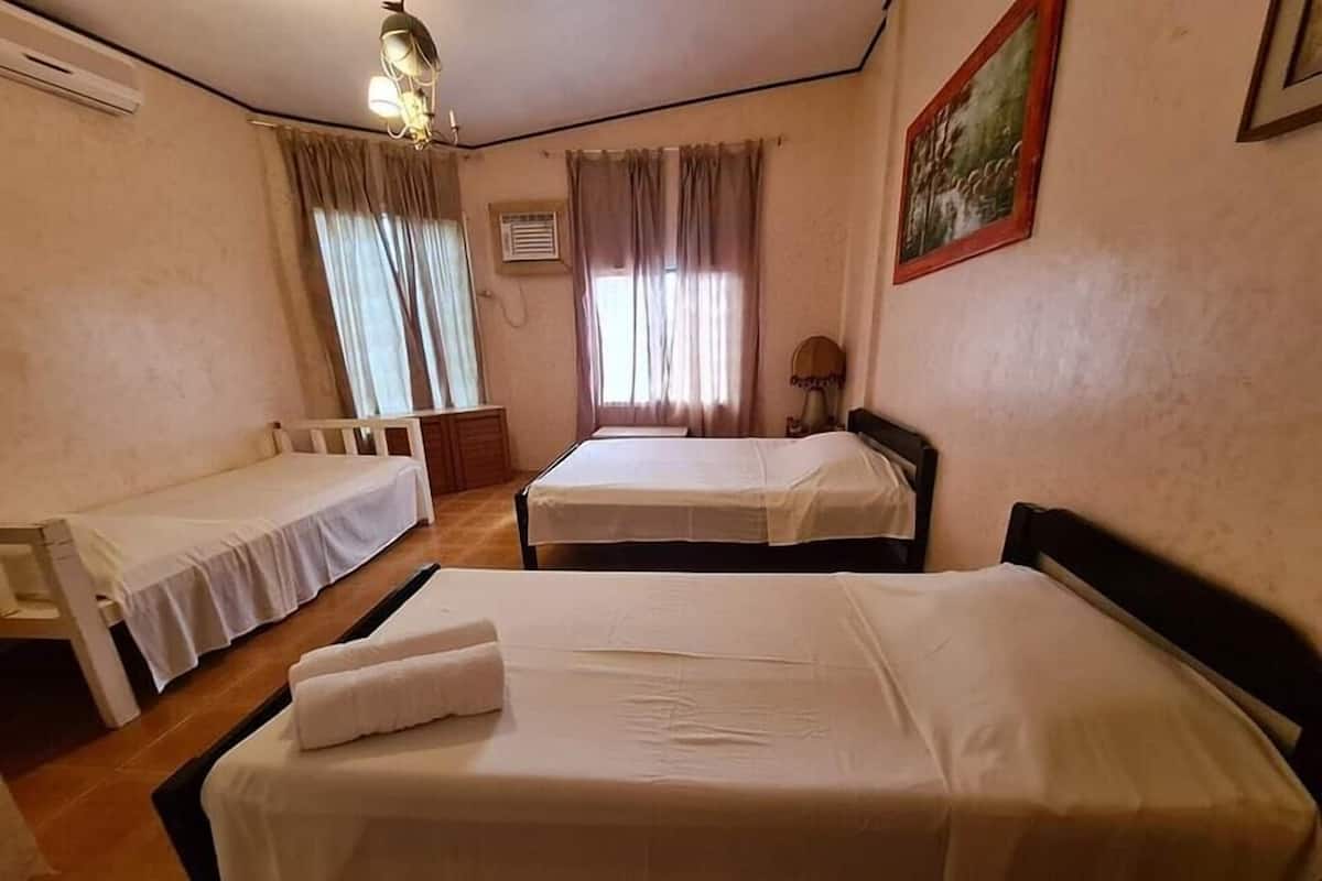 Quarto com duas camas de solteiro, uma mesinha e uma cadeira. Há uma janela com cortinas, um ar condicionado e um quadro na parede.