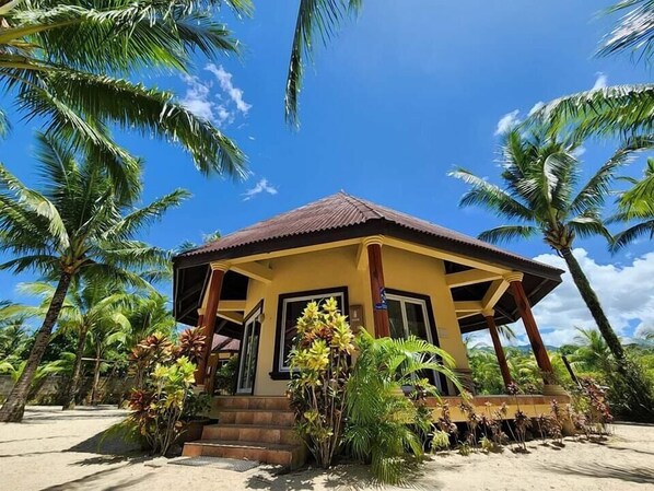 A small, single-story building with a brown roof and yellow walls, surrounded by palm trees and tropical plants. - Le Palme Beach Resort (Pandan)