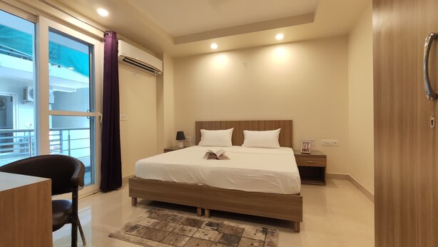 A hotel room with a bed, bedside table, lamp, and a view of the outside balcony. - Covie Gurgaon 70 (Gurugram)