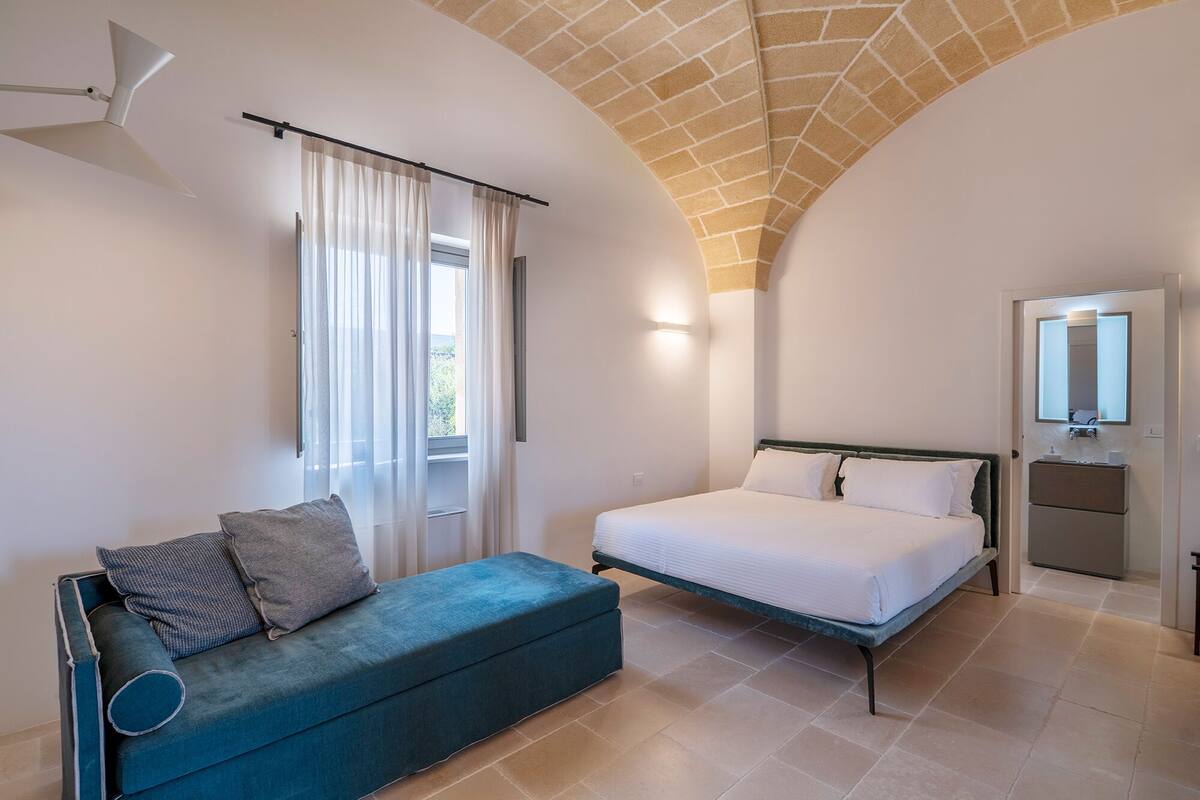 Un dormitorio con cama, sofá, ventana con cortinas, espejo y techo con diseño de ladrillo.
