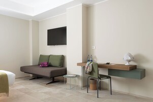 A modern room with a sofa, a small round table, a desk with a chair, a TV mounted on the wall, and a shelf with decorative items.