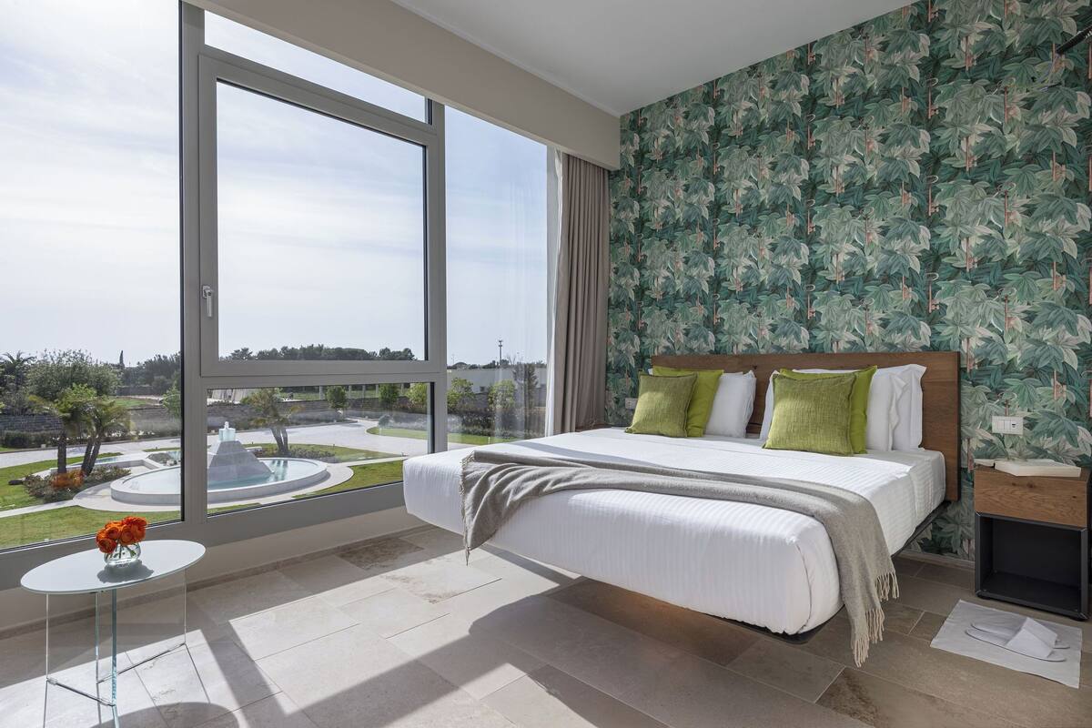Un dormitorio moderno con una cama grande, una mesita con flores y vistas a una fuente y vegetación al exterior.