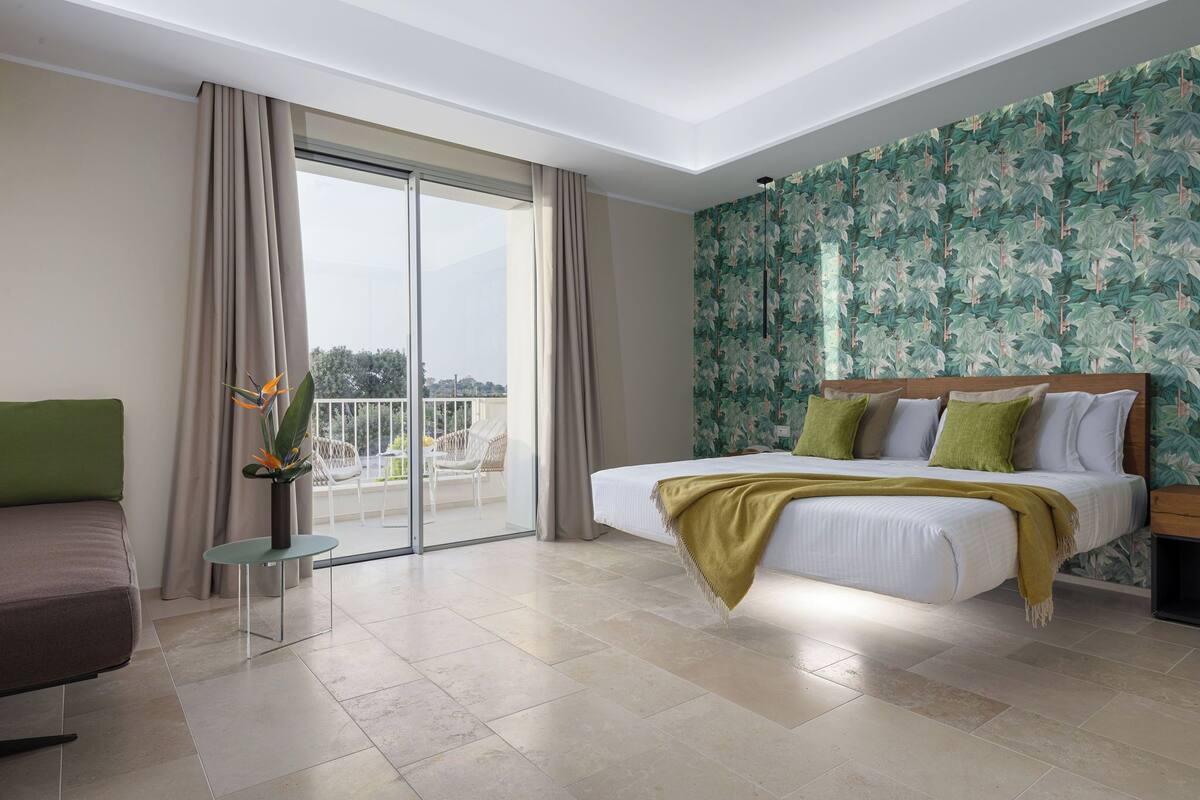 Una habitación de hotel moderna con una cama grande, un sofá, una mesita con un florero y un balcón con vistas.