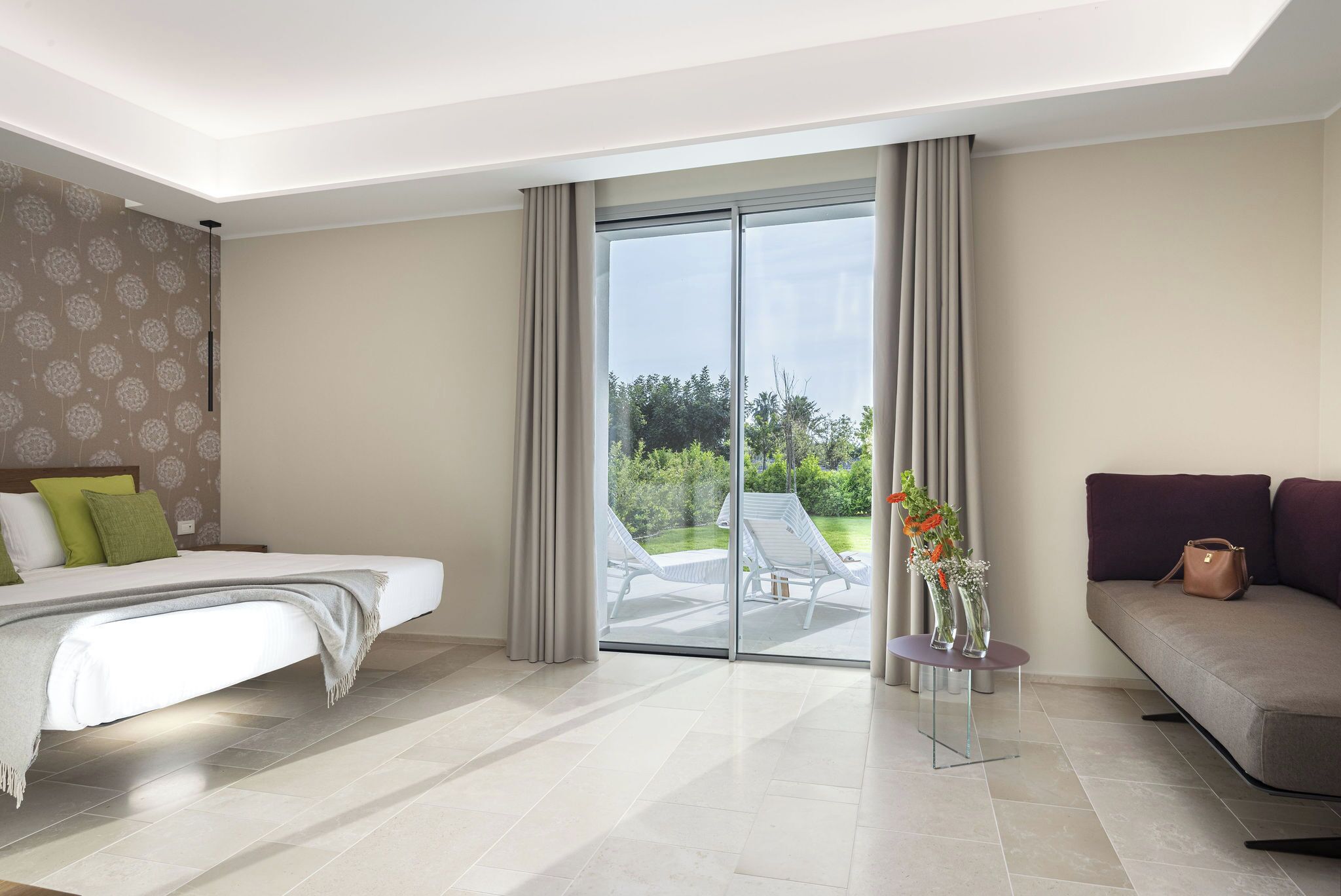 A modern hotel room with a bed, a sofa, a small table with vases, and a sliding glass door leading to an outdoor area with chairs.