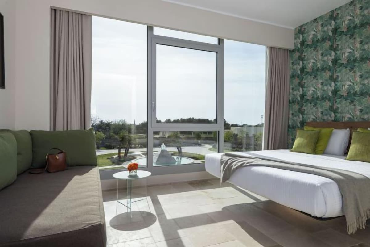 Una habitación de hotel moderna con un ventanal grande, una cama, un sofá y una mesita.