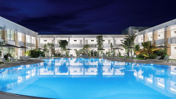 A large indoor swimming pool with lounge chairs and umbrellas, surrounded by a modern building with multiple floors and balconies. - Torre Assunta Hotel (Gallipoli)