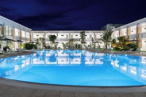 A large indoor swimming pool with lounge chairs and umbrellas, surrounded by a modern building with multiple floors and balconies.