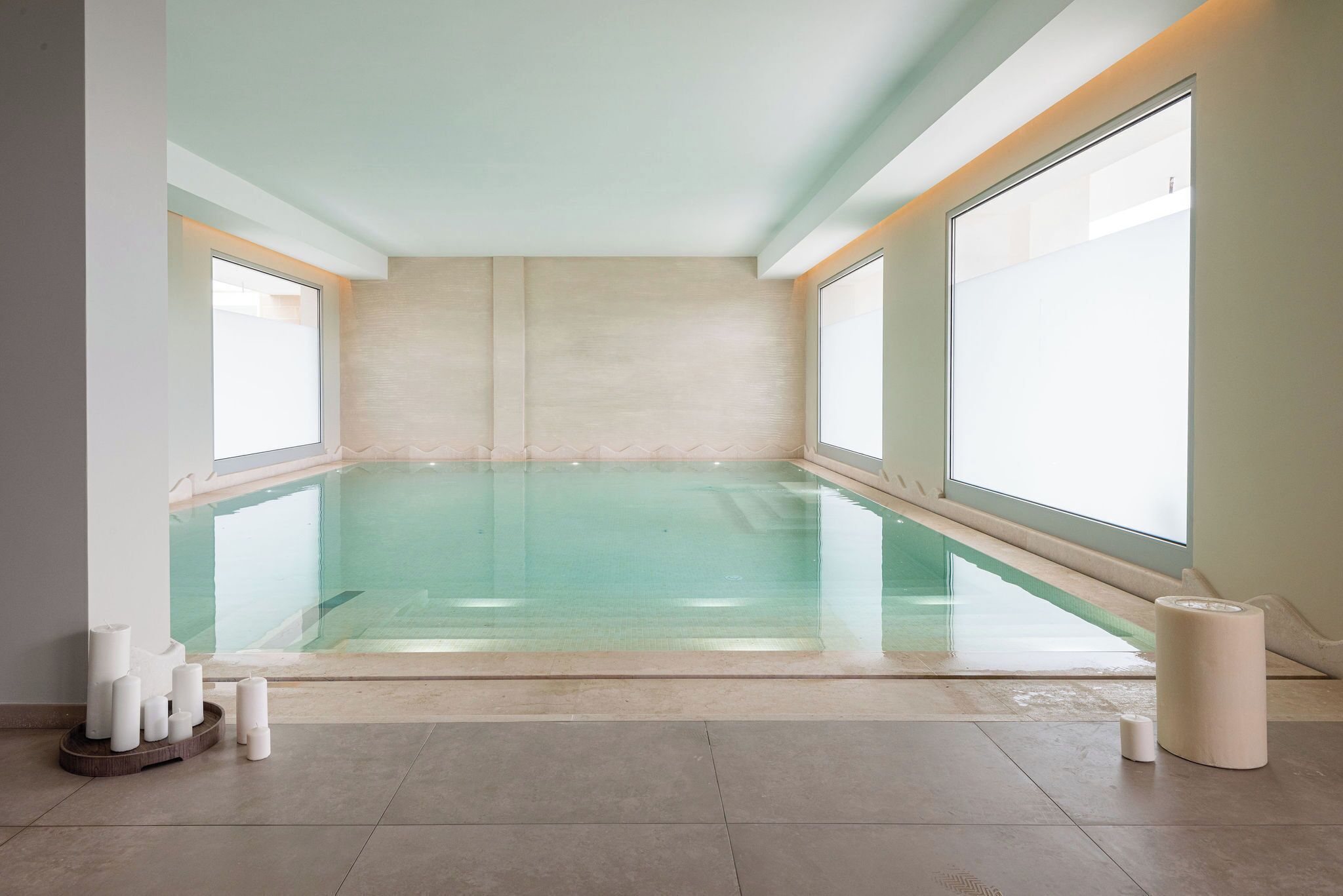 A modern indoor swimming pool with clear glass walls, large windows, and minimalistic decor.