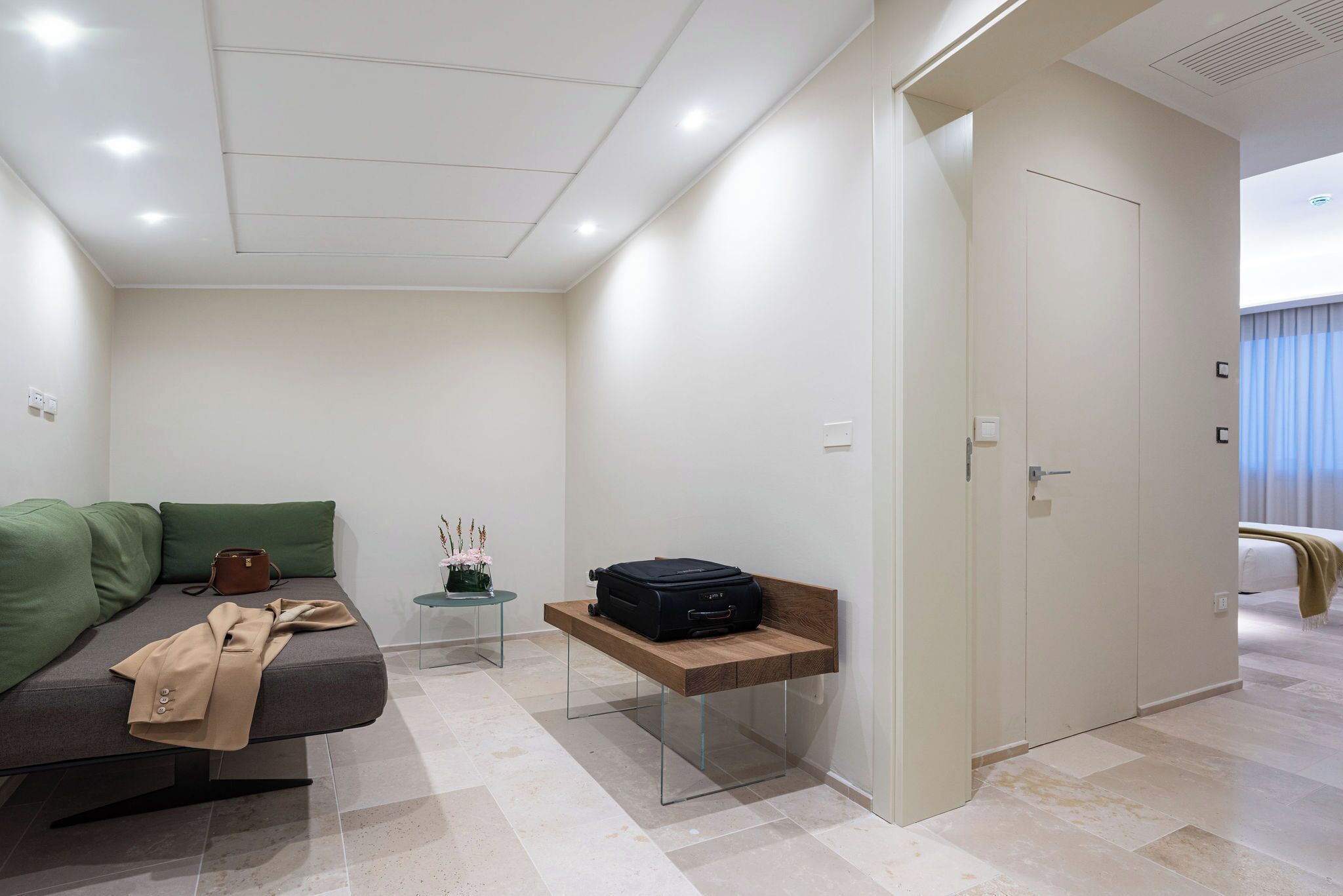 A modern hotel room with a sofa, a small table with a vase, a bench with a suitcase, and a wardrobe.