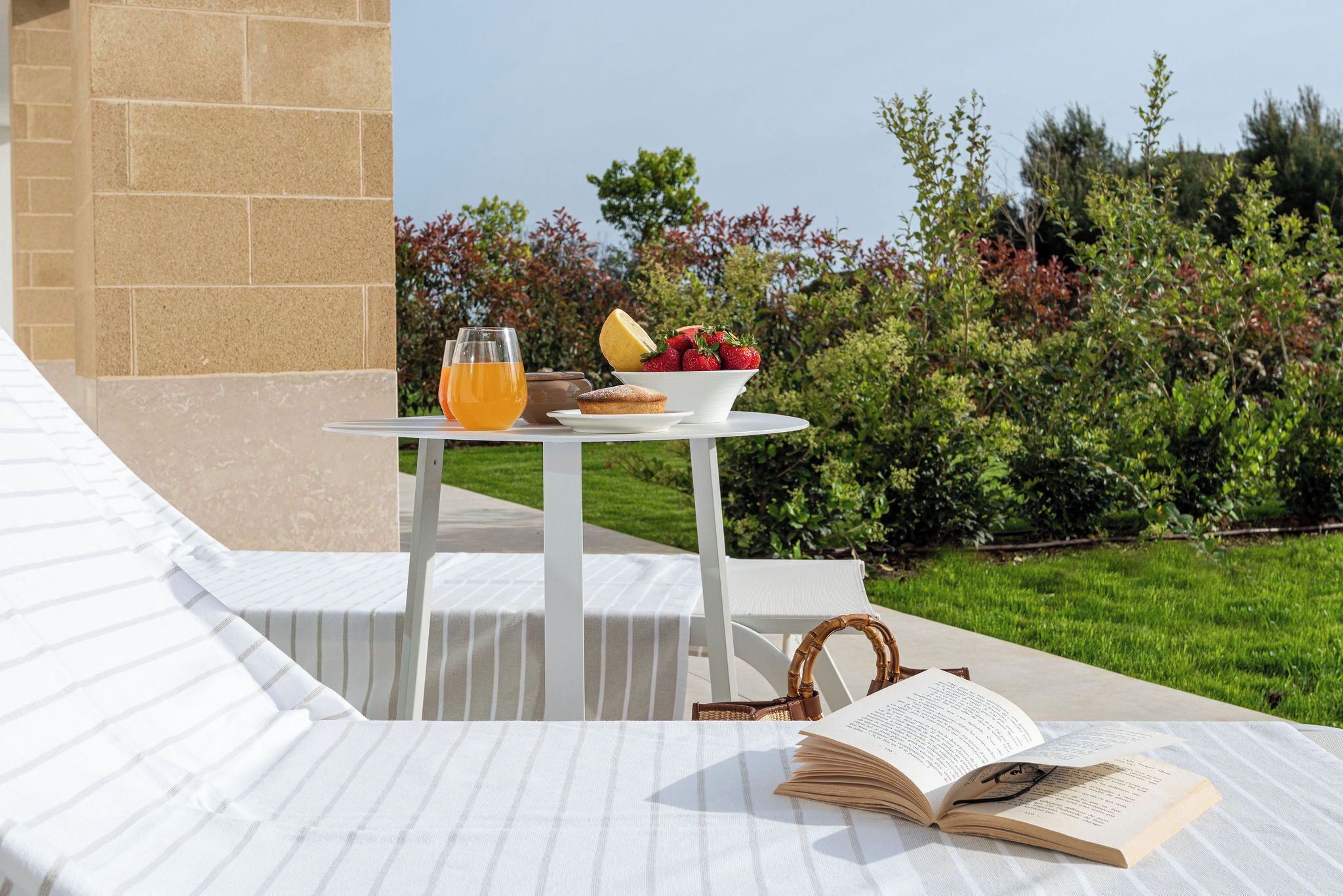 A table with a book, a glass of orange juice, and a bowl of fruit on a terrace with a view of greenery.