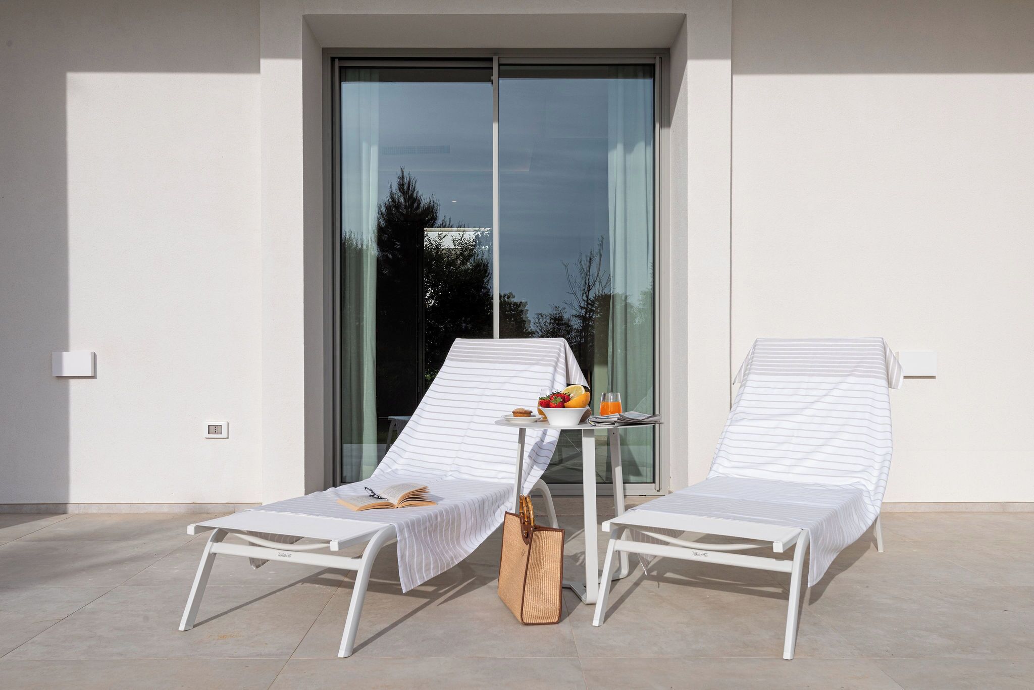 Two white lounge chairs with striped covers, a small table with a bowl of fruit and a drink, and a wicker bag on a balcony with a glass door and a view of trees.