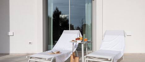 Two white lounge chairs with striped covers, a small table with a bowl of fruit and a drink, and a wicker bag on a balcony with a glass door and a view of trees.