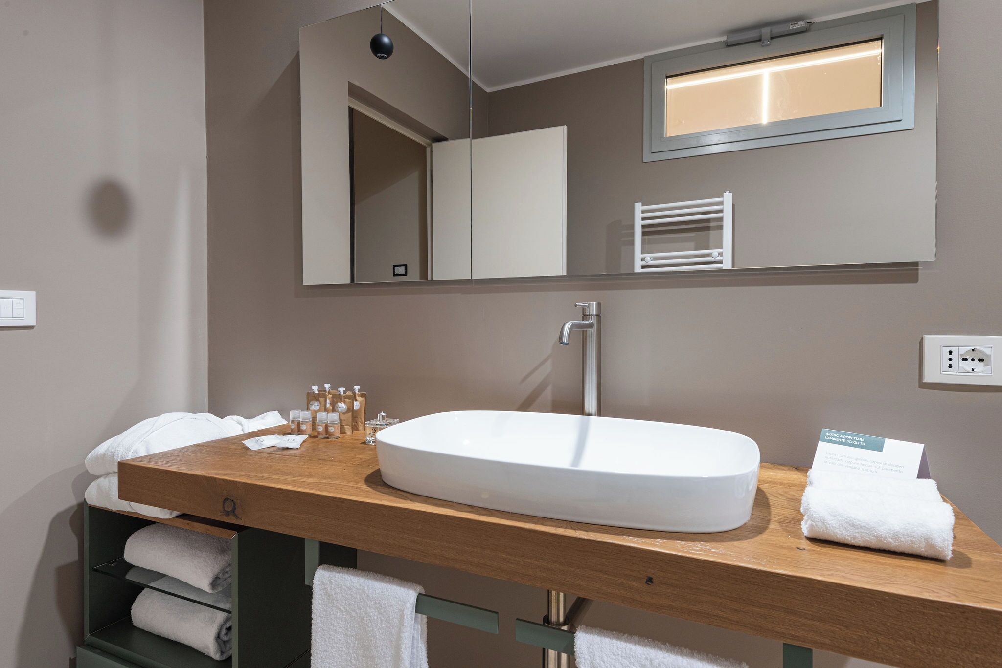 A modern bathroom with a wooden vanity, a white sink, and a large mirror. There are towels and toiletries on the vanity.