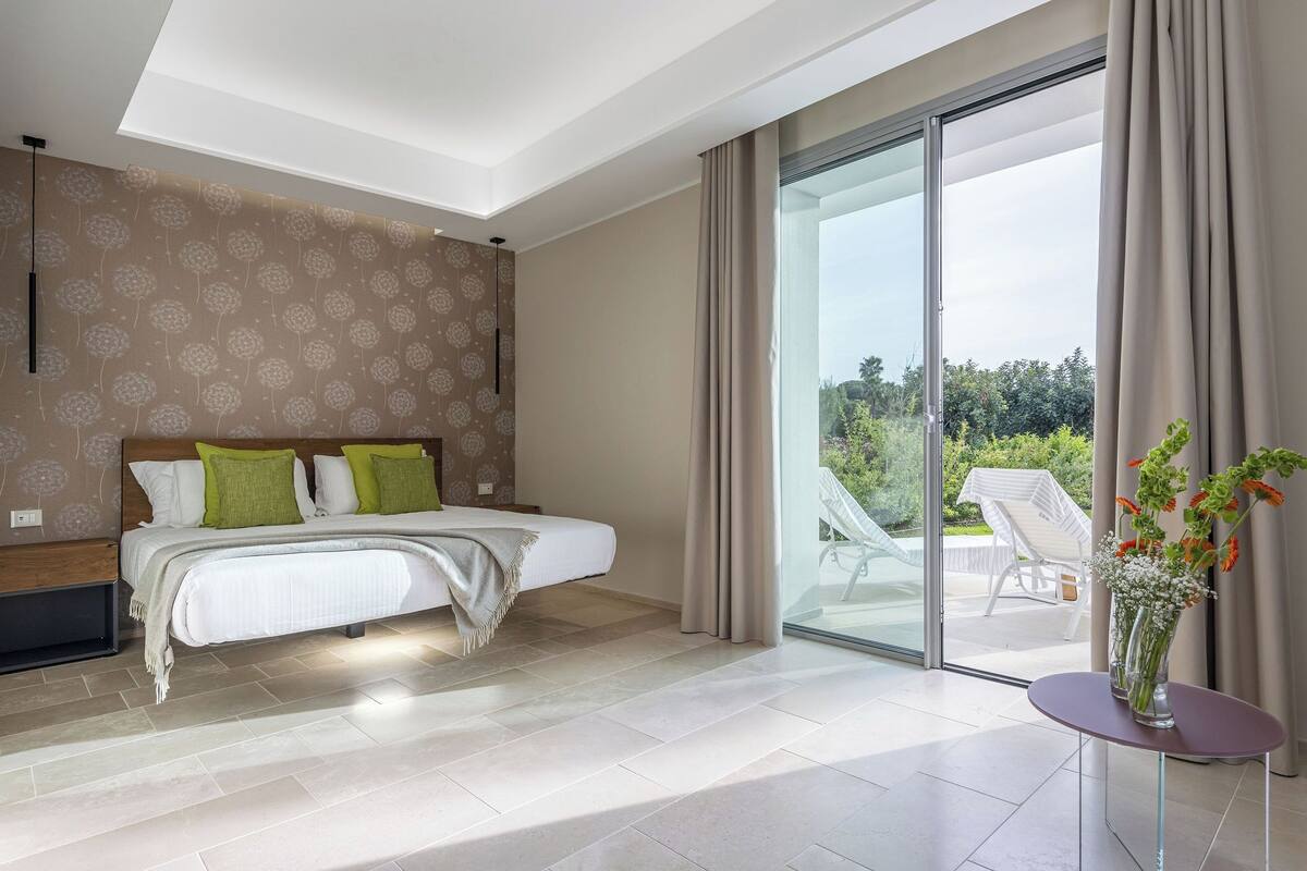 Un dormitorio moderno con una cama grande, una mesita de noche con un florero y un ventanal corredero que da a un espacio exterior.