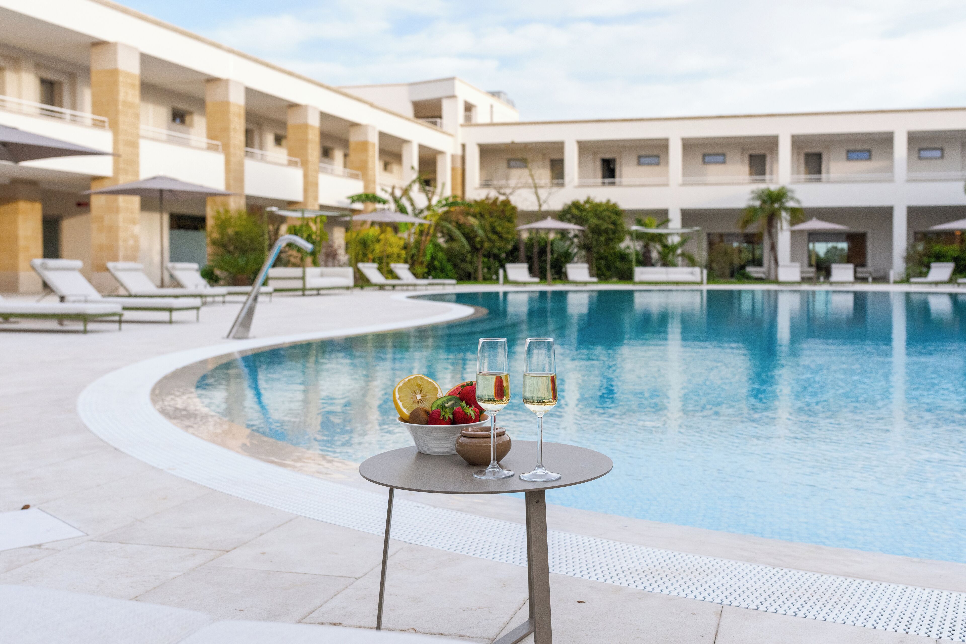 Un espace aménagé au bord de la piscine, avec des chaises longues, une table sur laquelle se trouve un saladier rempli de fruits et deux verres de vin, ainsi qu’une piscine.