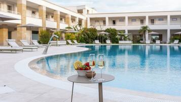 A poolside area with lounge chairs, a table with a bowl of fruit and two glasses of wine, and a pool.