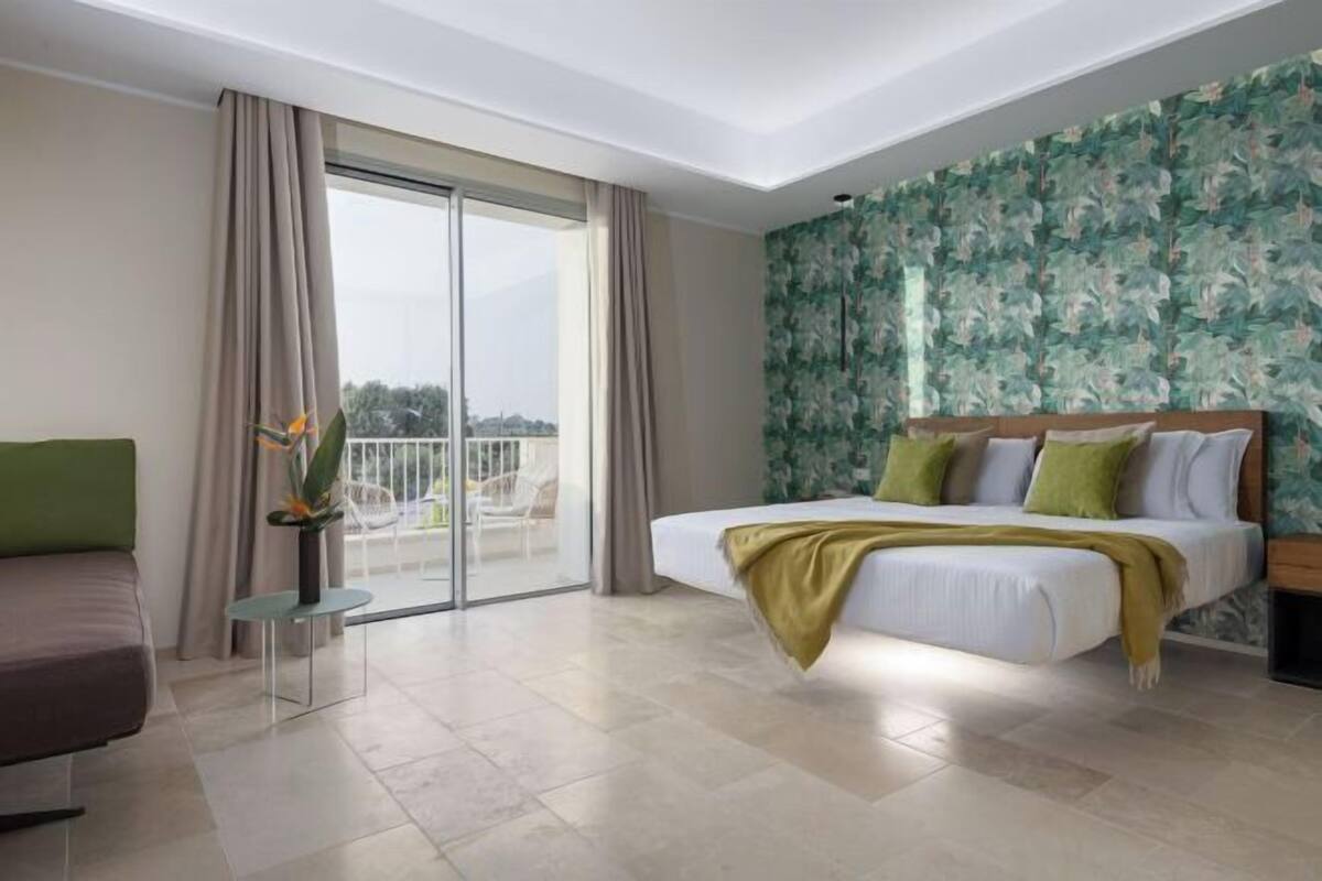 Una habitación de hotel con cama, sofá, una mesita con un florero y balcón con cortinas.